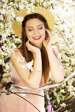Güzellik Portresi. Genç ve güzel uzun saçlı kız pembe elbise ve şapka ayakta çiçek arka plan üzerinde retro bisikleti ile gülümseyen.
