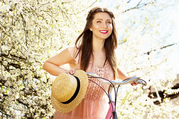 Blomma porträtt av anbud, leende ung och vacker långhårig flicka i rosa klänning cykla och att hålla hatten på blomma bakgrund. — Stockfoto