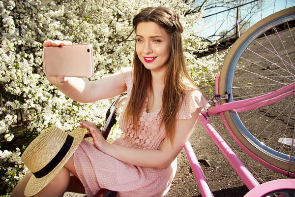 Rapariga Selfie. Retrato de sorrir jovem e bela menina de cabelos compridos em vestido rosa tomando selfie em seu telefone. Bicicleta retrô e flores no fundo . — Fotografia de Stock