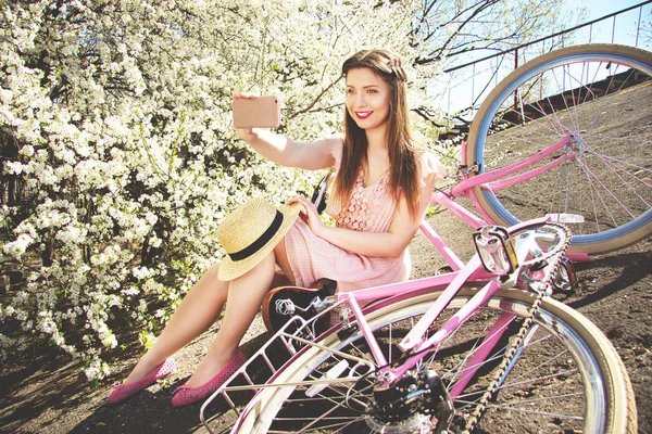 Selfie girl. Portrét usměvavé mladé a krásné dlouhé vlasy dívka v růžových šatech pořizování selfie na telefonu. Retro kolo a květiny na pozadí. — Stock fotografie