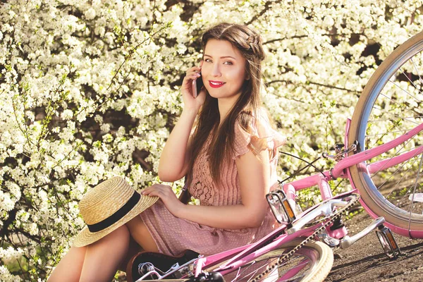 Un poco de conversación. Retrato de joven y hermosa chica de pelo largo en vestido rosa hablando por teléfono y sentado con bicicleta retro sobre fondo de flores . — Foto de Stock