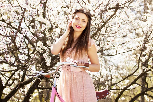 Retrato de beleza da menina macia, atraente e jovem de cabelos longos em vestido rosa de pé com bicicleta retro no fundo da flor . — Fotografia de Stock