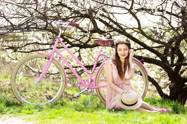 Hermosa naturaleza primaveral. Atractiva joven y hermosa chica de pelo largo en vestido rosa sentado con bicicleta retro en la hierba verde. Fondo de flores . — Foto de Stock