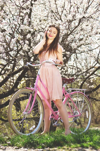 우아한 여자입니다. 녹색 잔디에 핑크 레트로 자전거 서 있는 핑크 드레스에 긴 머리 소녀 아름 답 고 젊은 꽃 배경. — 스톡 사진