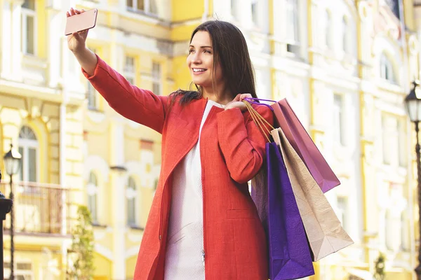 Hora de selfie. Retrato de mujer joven y atractiva en abrigo rojo tomando una selfie en su teléfono con bolsas de compras . — Foto de Stock