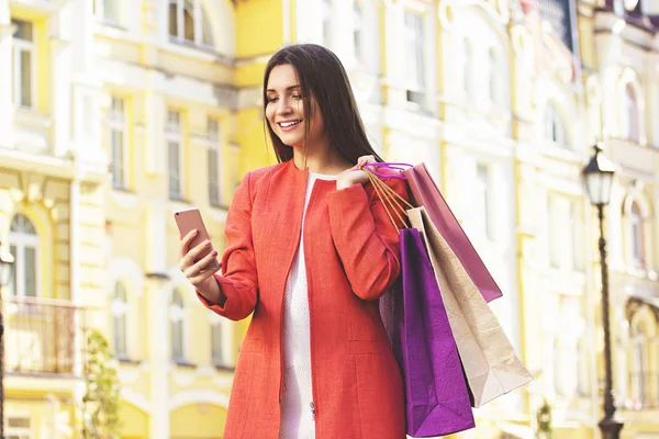 Mensajes al aire libre. Retrato de mujer hermosa y joven en abrigo rojo usando su teléfono mientras sostiene bolsas de compras . —  Fotos de Stock