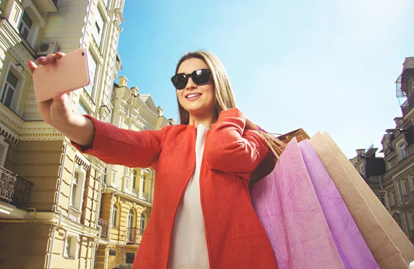 Selfie com compras. Retrato de mulher bonita sorridente em casaco vermelho e óculos de sol tirando uma selfie com sacos de compras . — Fotografia de Stock