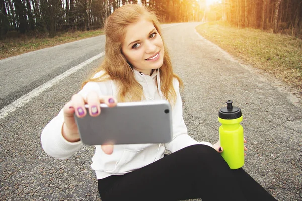 Selfie idő. A csinos, fiatal redhaired álló lány vesz egy selfie ülve a park road sport. — Stock Fotó
