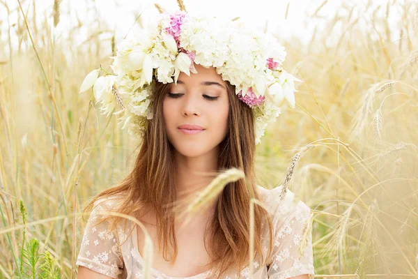 Beleza natural. Retrato de bela menina vestida de vestido branco e flores na cabeça está sentado no campo de trigo amarelo . — Fotografia de Stock