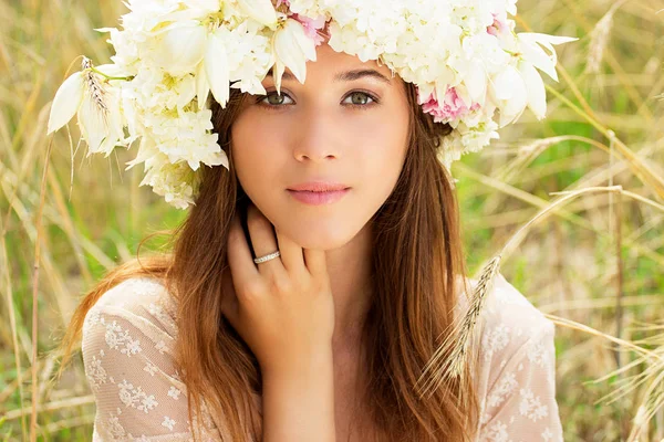 Retrato de beleza. Fechar-se de linda jovem mulher vestida de vestido branco e flores em sua cabeça está posando no campo de trigo amarelo . — Fotografia de Stock