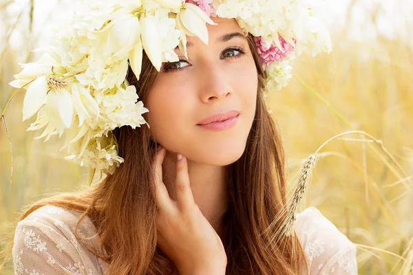 Beauty portrait. Closeup of gorgeous young woman dressed in white dress and flowers on her head is posing in yellow wheat field. — Stock Photo, Image