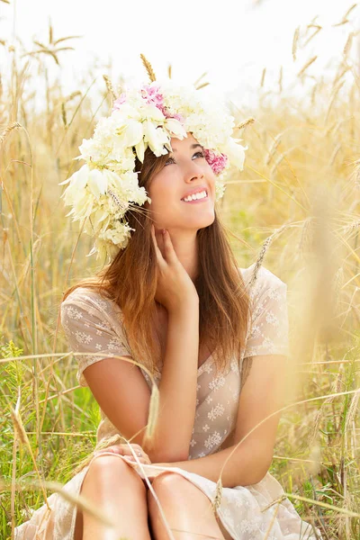 자연의 기쁨입니다. 젊은 여성이 그녀의 머리에 꽃과 흰색 드레스에서를 입고 노란 밀밭에 앉아 아름 다운 미소의 초상화. — 스톡 사진