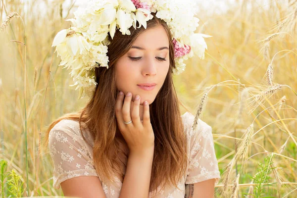 Perfekt skönhet. Porträtt av attraktiv ung kvinna klädd i vit klänning och blommor på hennes huvud vidrör hennes ansikte medan du sitter i den gula vetefält. — Stockfoto