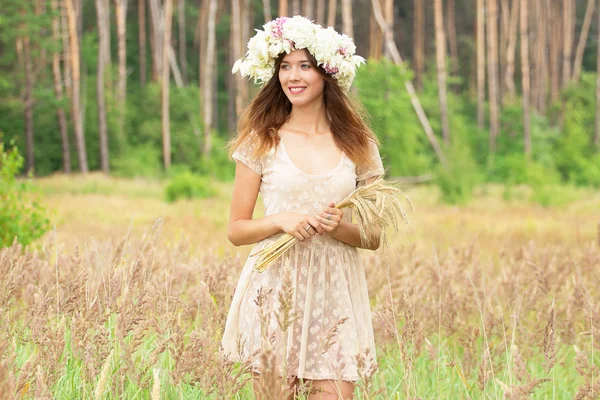 Feliz com a natureza. Retrato de jovem extremamente bonita em vestido branco e flores na cabeça mantendo o trigo em suas mãos, enquanto em pé no campo de trigo amarelo . — Fotografia de Stock