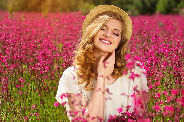 Cieszyć się życiem. Portret przepiękny młoda redhead dziewczyna ubrana w białą sukienkę i kapelusz pozuje do kamery siedząc na łące kwiat. — Zdjęcie stockowe