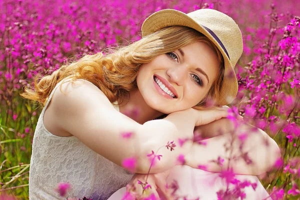 Gott humör. Porträtt av attraktiv ung rödhårig flicka klädd i vit klänning och hatt poserar i kameran när du sitter i blomma äng. — Stockfoto