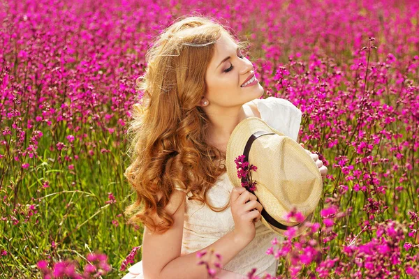 시골의 아름다움입니다. 하얀 드레스를 입은 젊고 매력적인 빨간 머리 여자의 꽃 초원에 서 있는 동안 카메라에 포즈. — 스톡 사진