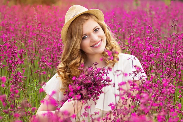 행복 한 아름다움입니다. 하얀 드레스 입은 아름 다운 젊은 빨간 머리 여자의 초상화와 모자는 꽃 시골 풀밭에 앉아있는 동안 카메라에 포즈. — 스톡 사진