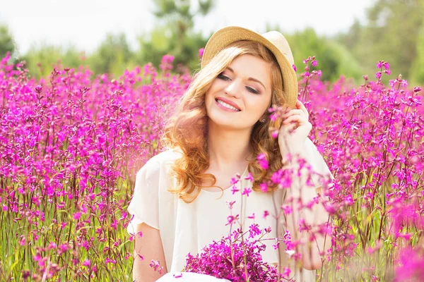 Feliz beleza. Retrato de bela menina ruiva vestida de vestido branco e chapéu está posando para a câmera enquanto sentado no prado do campo de flores . — Fotografia de Stock