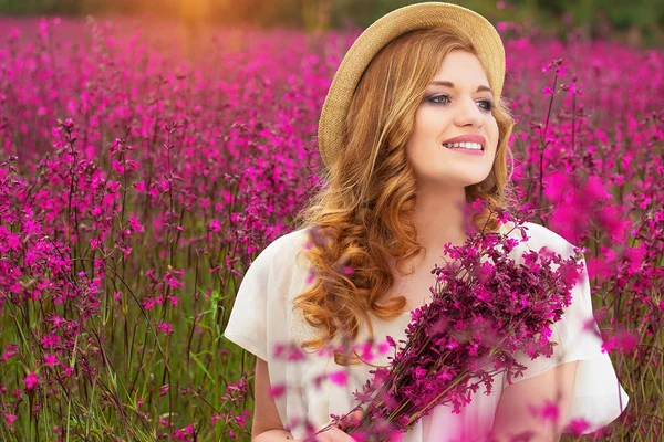 Ciesząc się charakter. Portret piękne młode Rude Girl ubrany w kapelusz i biała sukienka jest utrzymanie kwiaty podczas pozowanie w kwiat wsi łąka. — Zdjęcie stockowe