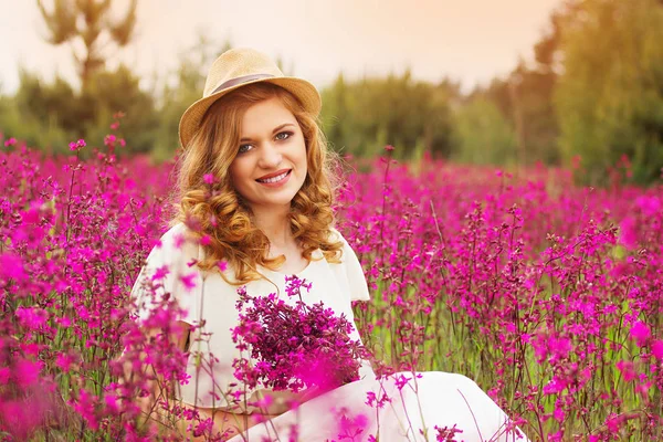 행복은 가까이 이다. 하얀 드레스 입은 아름 다운 젊은 여자의 초상화와 모자는 꽃 초원에 앉아있는 동안 그녀의 손에 꽃을 유지. — 스톡 사진