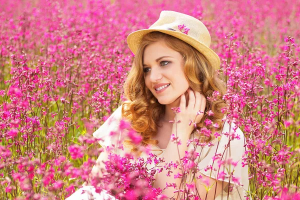 Feliz como uma criança. Retrato de bela menina ruiva vestida de chapéu e vestido branco está mantendo flores enquanto posando no prado do campo de flores . — Fotografia de Stock