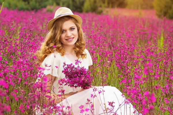 A felicidade está perto de ti. Retrato de bela menina vestida de vestido branco e chapéu está mantendo flores em suas mãos enquanto sentado no prado da flor . — Fotografia de Stock