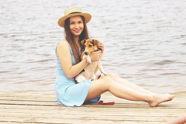 Happy owner. Horizontal shot of beautiful and young woman in dress holding little dog in her hands and smiling while sitting on wooden pier. — Stock Photo, Image