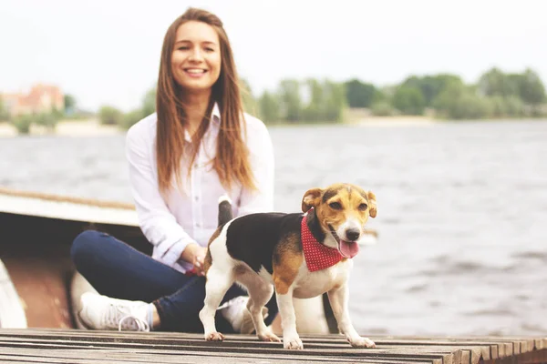 Divertirmo-nos juntos. Horizontal tiro na bela jovem mulher em desgaste casual está olhando para a câmera e sorrindo enquanto sentado no cais com seu lindo cão . — Fotografia de Stock