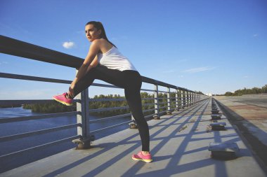 Eğitim için iyi bir gün. Kendine güvenen, güzel ve genç kadın holding bacak giyim ve uzak zemin üzerine mavi gökyüzü ile köprüde germe yaparken seyir spor yatay çekim.