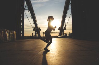 O akşam koşu seviyor. Arka plan üzerinde endüstriyel manzaralı günbatımı sırasında köprüde çalışan yan görünümü atış genç güzel kadın spor giyim.