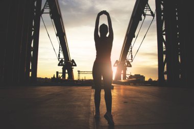 Koşu önce ısınma. Silhouette sportif kadın silah kaldırdı tutmak ve akşam güneş ışığı ile köprü üzerinde duran ve endüstriyel görünümü arka planı süre germe yapıyor.