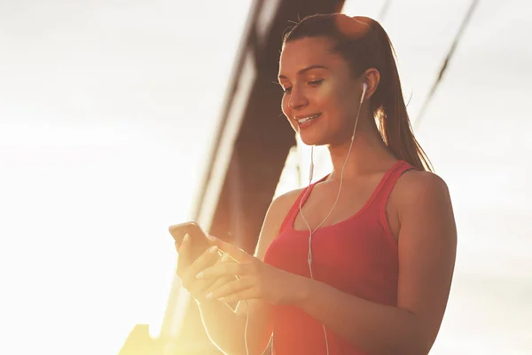 Nagy nap a képzés. Sportruházat gazdaság szúró telefon és látszó-on ez a híd a háttérben esti napfény állva vonzó és fiatal vidám nő portréja. — Stock Fotó