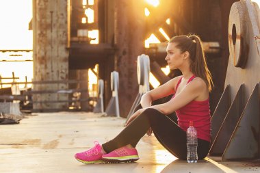 Akşam dürtmek sonra dinlenme. Çekici genç kadın spor yan görünüm giymek uzakta akşam güneş ışığı ve arka plan üzerinde endüstriyel görünümü ile köprü üzerinde otururken arıyorsunuz.