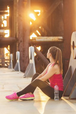 Bir fren al. Güzel genç kadın spor yan görünüm giymek uzakta akşam güneş ışığı ve arka plan üzerinde endüstriyel görünümü ile köprü üzerinde otururken arıyorsunuz.