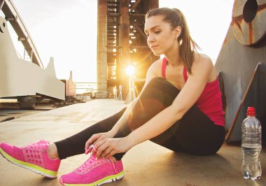 Küçük fren akşam koşu sırasında. Güzel ve genç spor kadın arka plan üzerinde akşam güneş ışığı ile köprü üzerinde otururken spor ayakkabılarını üzerinde ayakkabı bağcığı bağlama.