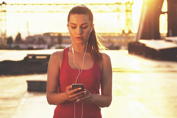 Gadgets für den Sport. Porträt einer selbstbewussten schönen Frau in Sportkleidung, die ihr Smartphone benutzt, während sie auf einer Brücke mit Abendsonne im Hintergrund steht. — Stockfoto