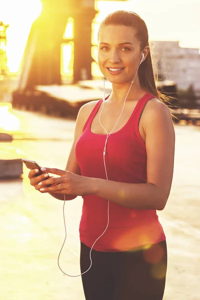 Motivationsmusik. attraktive junge Sportlerin blickt in die Kamera und lächelt, während sie während des Trainings auf einer Brücke mit Sonnenlicht und Stadtansichten im Hintergrund telefoniert. — Stockfoto