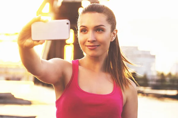 Sie liebt Selfie. Porträt einer jungen und attraktiven Frau in Sportkleidung, die auf ihr Handy schaut und lächelt, während sie während des Outdoor-Trainings ein Selfie mit ihrem Handy macht. — Stockfoto