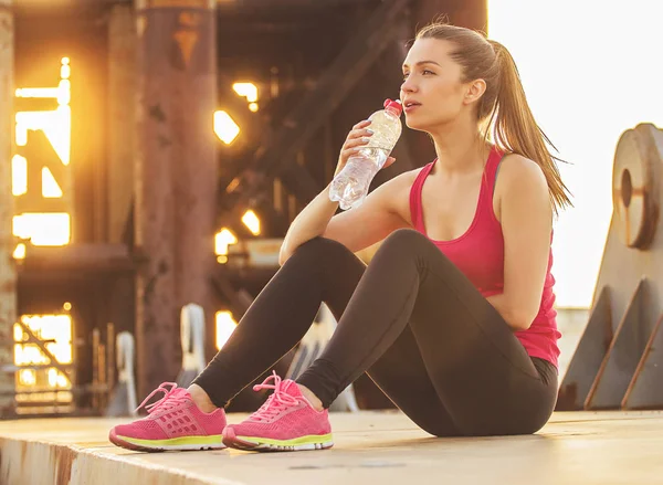 Tartása a víz egyensúlyát. Vízszintes lövés a gyönyörű fiatal nő, a sport viselni keres el és ivóvíz után jogging ülve a hídon. — Stock Fotó