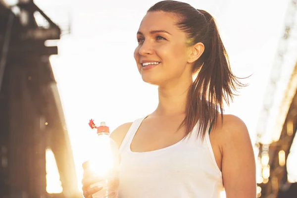 Fröhliche Fitness-Mädchen. Niedrigwinkel-Ansicht der jungen attraktiven Sportlerin, die wegschaut und lächelt, während sie Wasser auf der Brücke trinkt, mit Sonnenlicht im Hintergrund. — Stockfoto