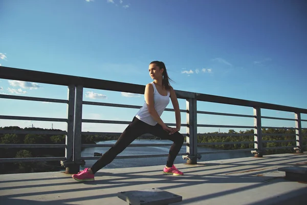 Bemelegítés, edzés előtt. Vízszintes lövés a gyönyörű fiatal nő, a sport viselni keres el eközben nyúlik a hídon a kék ég, a háttérben. — Stock Fotó