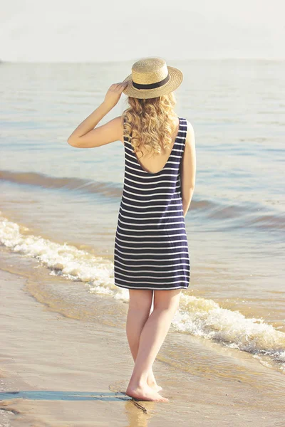 Beauté romantique. Vue arrière de belle et jeune femme blonde en robe ajustant le chapeau sur la tête tout en marchant sur la plage de la mer . — Photo