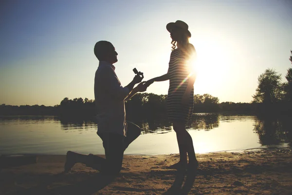 Wirst du mich heiraten? Silhouette eines jungen Mannes, der auf den Knien bleibt und seinem hübschen Mädchen am Strand mit Abendsonne auf dem Hintergrund einen Heiratsantrag macht. — Stockfoto