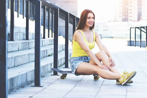 Сідайте на гальма. Горизонтальний знімок молодої і привабливої жінки в хіпстерському одязі дивиться і посміхається, сидячи на скейтборді біля сходів стадіону . — стокове фото