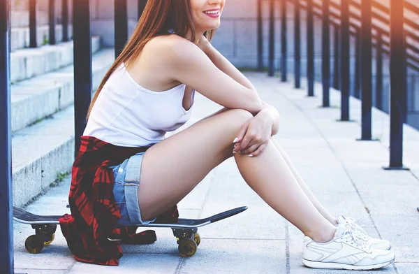 Relaks po skateboarding day. Ujęcie poziome strony piękna młoda kobieta w hipster nosić odwracając i uśmiechając się siedząc na deskorolce. — Zdjęcie stockowe