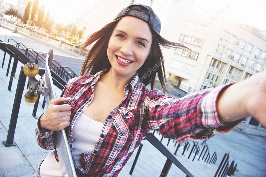 Happy moments must be saved! Cheerful, young and beautiful hipster woman in shirt and cap holding camera and taking selfie while keeping skateboard in another hand.