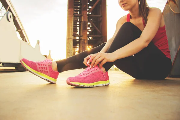 Ruhe nach dem abendlichen Joggen. attraktive und junge Sportlerin, die Schnürsenkel an ihre Turnschuhe bindet, während sie auf einer Brücke sitzt und Abendsonne auf dem Hintergrund. — Stockfoto