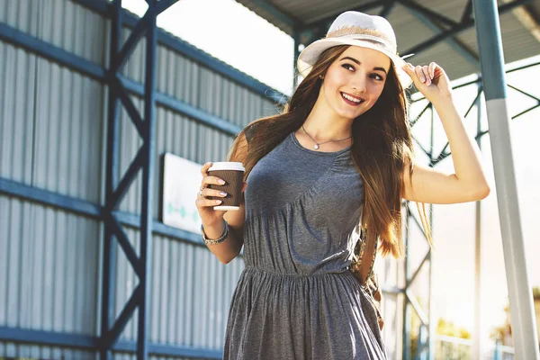 Веселий турист. Портрет красивої молодої жінки в одязі і капелюсі, що регулює капелюх на голові і посміхається під час пиття кави на зупинці поїзда . — стокове фото