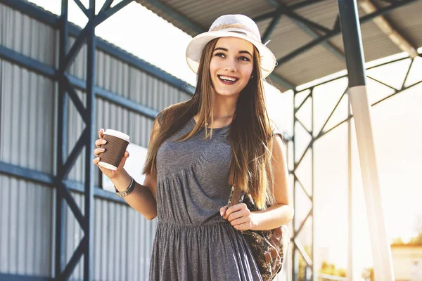 Frein à café. Portrait de jolie jeune femme en robe et chapeau regardant loin et souriant tout en buvant du café sur l'arrêt de train urbain. Voyage fille . — Photo
